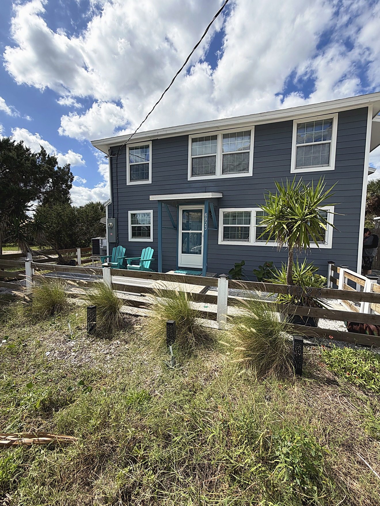 Front exterior of a two-story coastal rental home with turquoise chairs on the front deck, photographed for a short-term rental listing