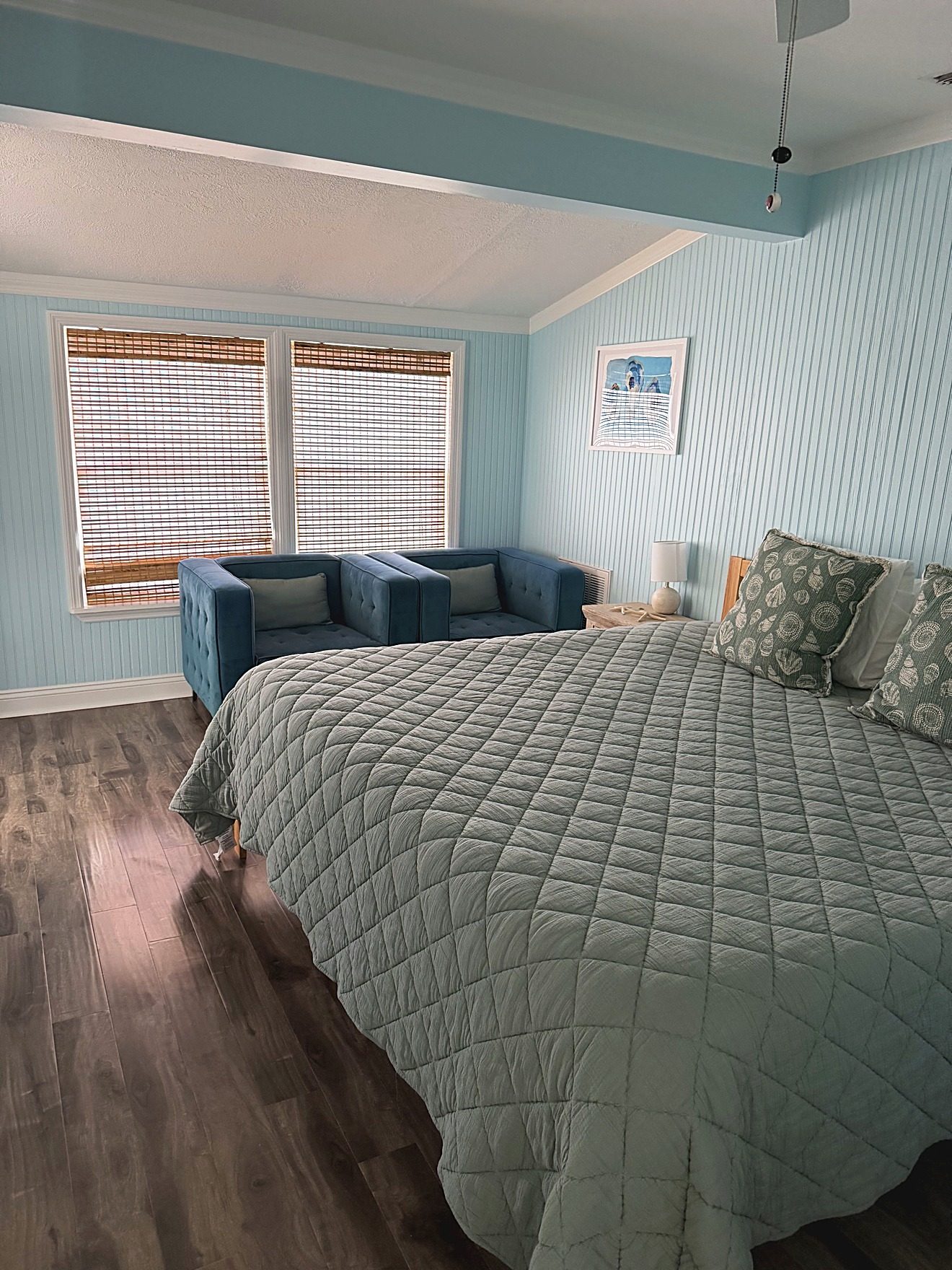 Primary bedroom with a quilted bedspread, two blue armchairs, and bamboo blinds in a coastal rental home