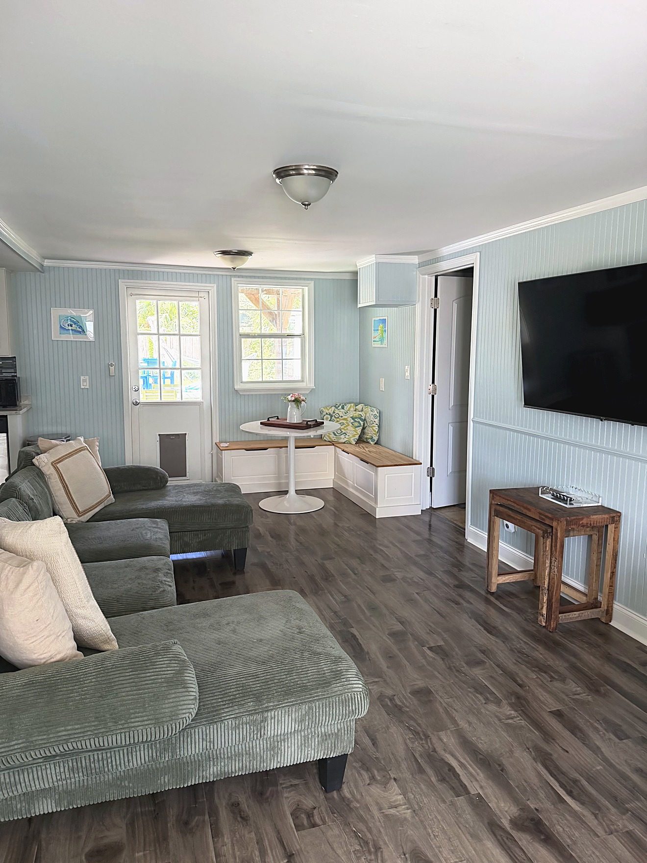Living room of a coastal rental home with a green sectional, built-in dining bench, and wall-mounted TV, photographed for a listing