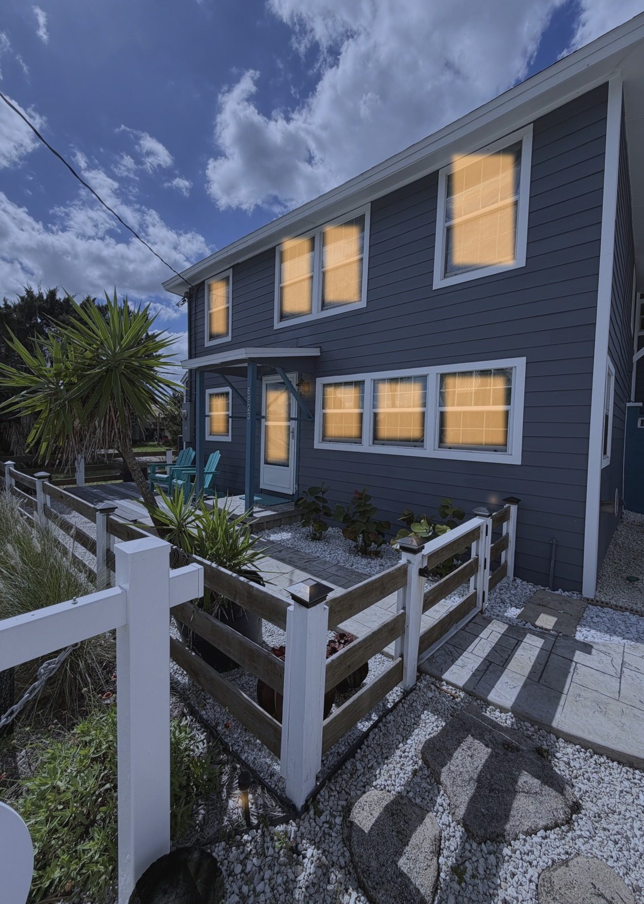 Angled twilight view of a two-story coastal home with warm glowing windows and a fenced front deck