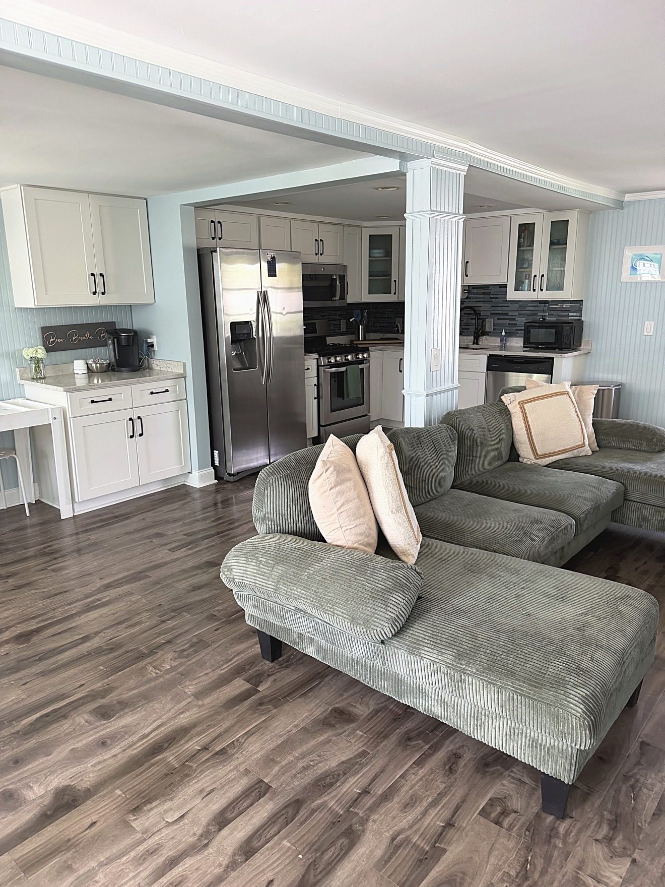 Open living room and kitchen of a coastal rental home with a green sectional, white cabinets, and stainless appliances