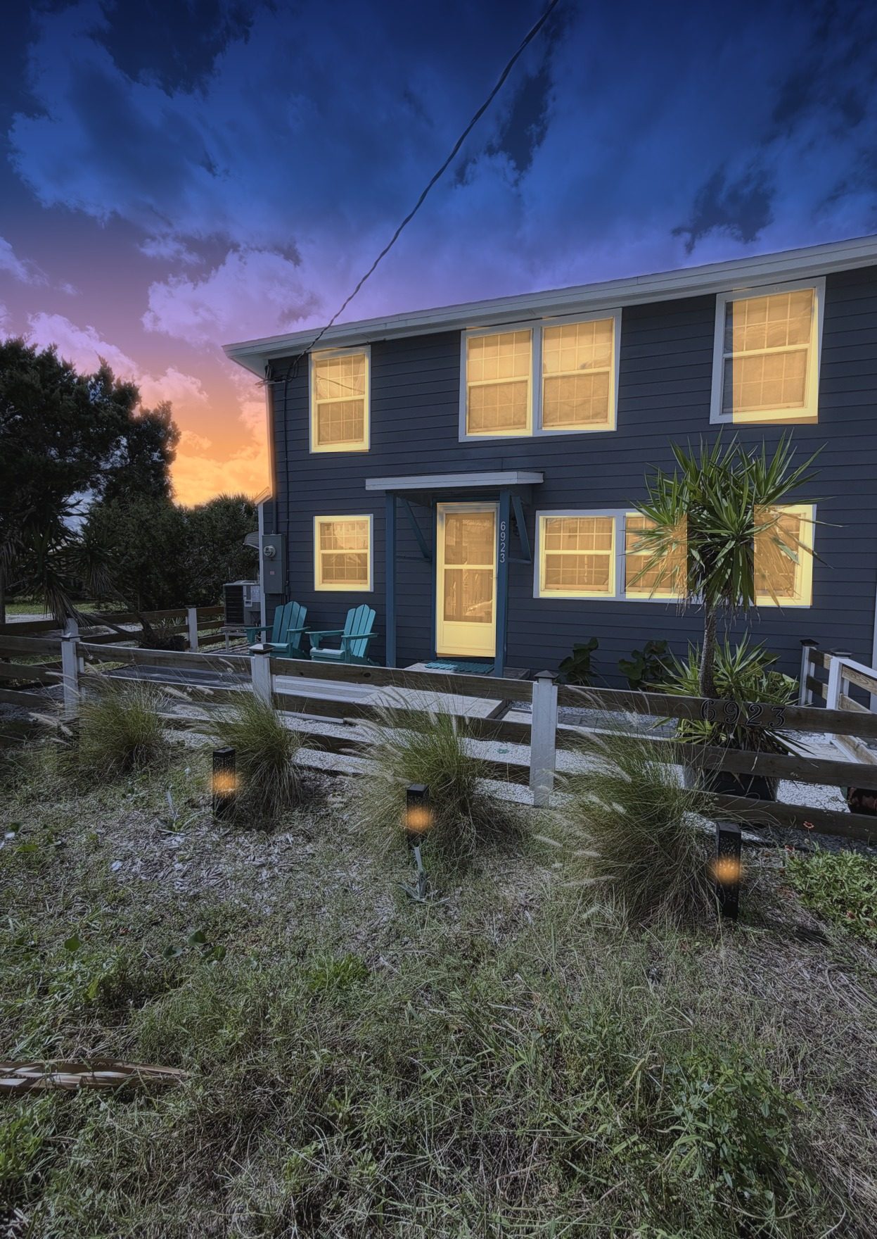 Twilight front exterior of a two-story coastal home with warm glowing windows under a sunset sky